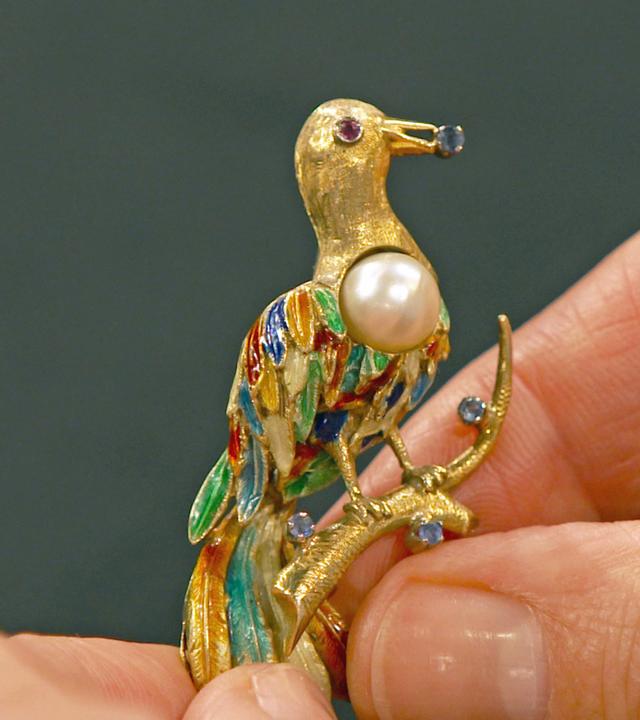 Ein goldene Brosche in Form eines Paradiesvogels mit einer Perle und kleinen, bunten Edelsteinen