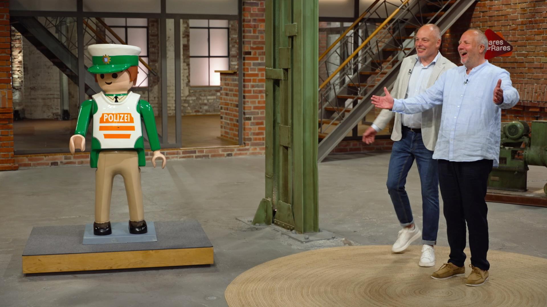 Im Händlerraum stehen rechts zwei Verkäufer und links eine menschengroßer Playmobil-Polizist.