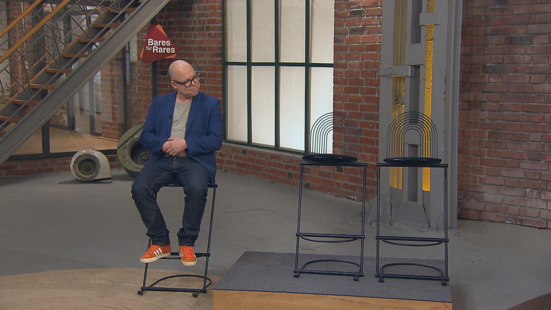 Händler Benjamin Leo Leo sitzt auf einem Designer-Barhocker. Daneben stehen zwei weitere Barhocker.