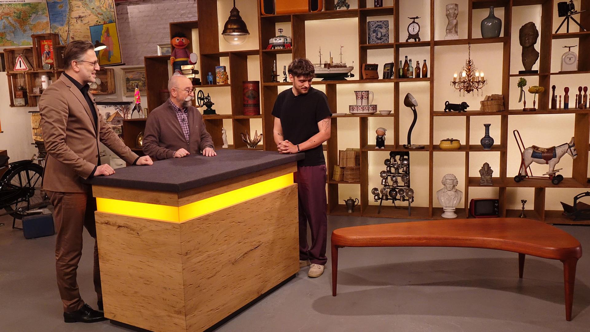 Experte Detlev Kümmel begutachtet einen Couchtisch. Neben ihm am Expertentisch stehen Horst Lichter und der Verkäufer des Tischs.