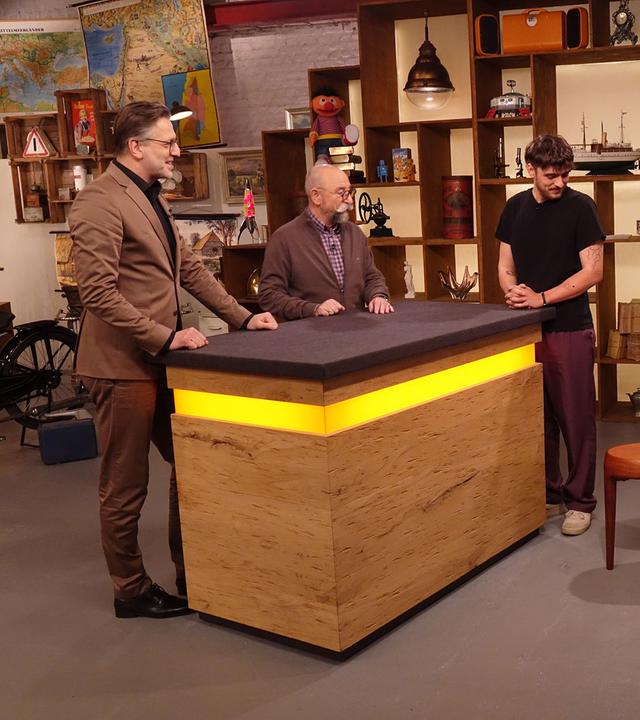 Experte Detlev Kümmel begutachtet einen Couchtisch. Neben ihm am Expertentisch stehen Horst Lichter und der Verkäufer des Tischs.