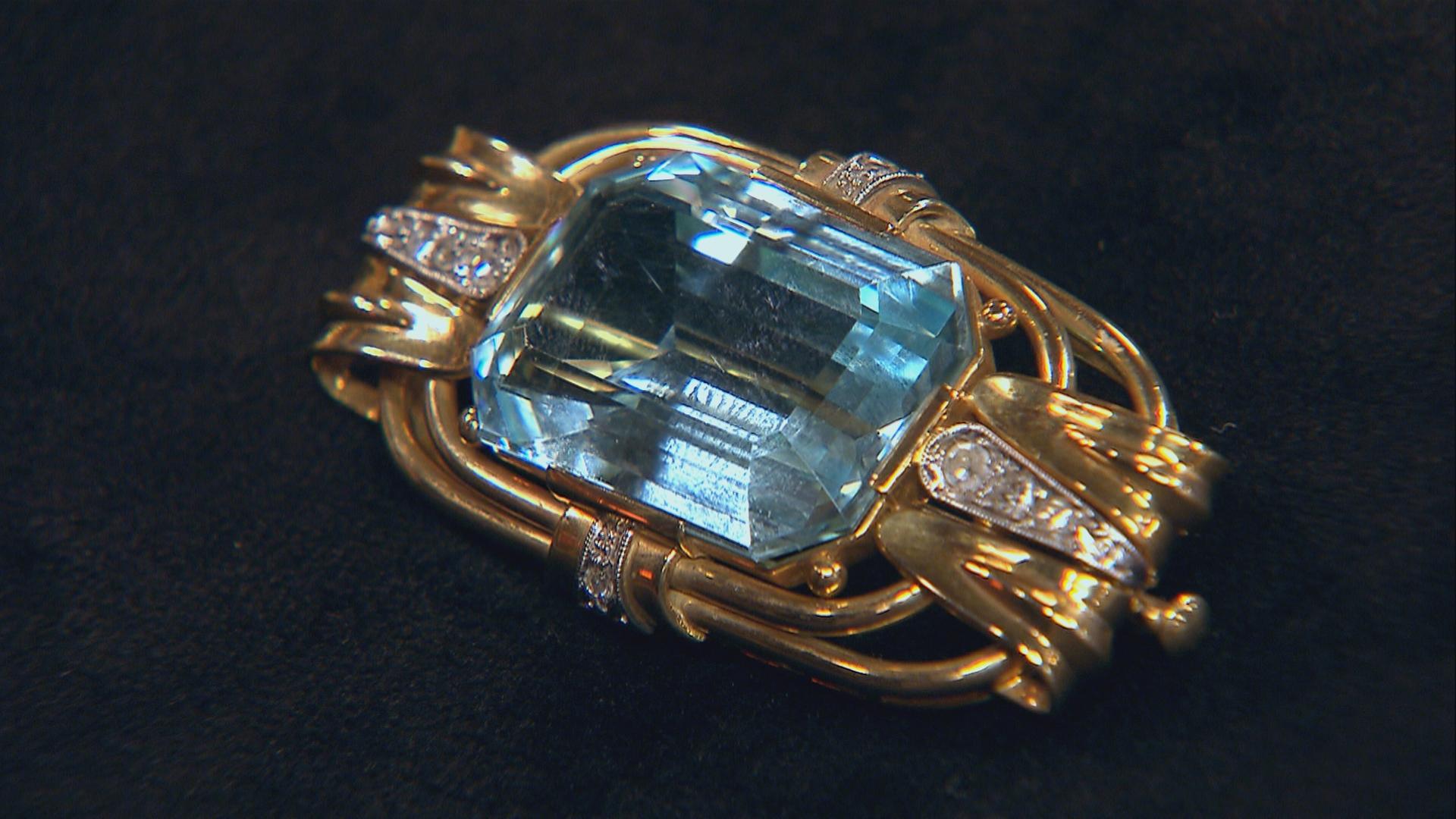 Eine goldene Brosche liegt auf schwarzem Grund. Der große Aquamarin in der Mitte der Brosche ist Rechteckig geschliffen.