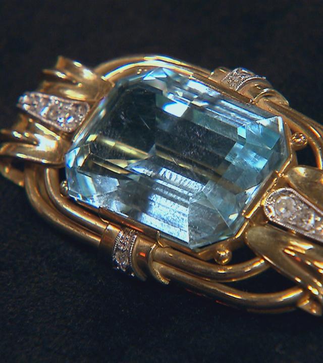 Eine goldene Brosche liegt auf schwarzem Grund. Der große Aquamarin in der Mitte der Brosche ist Rechteckig geschliffen.