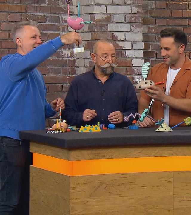 Die Händler Jan Cizek und David Suppes stehen mit Moderator Horst Lichter im Händlerraum und spielen mit Holzspielzeug.