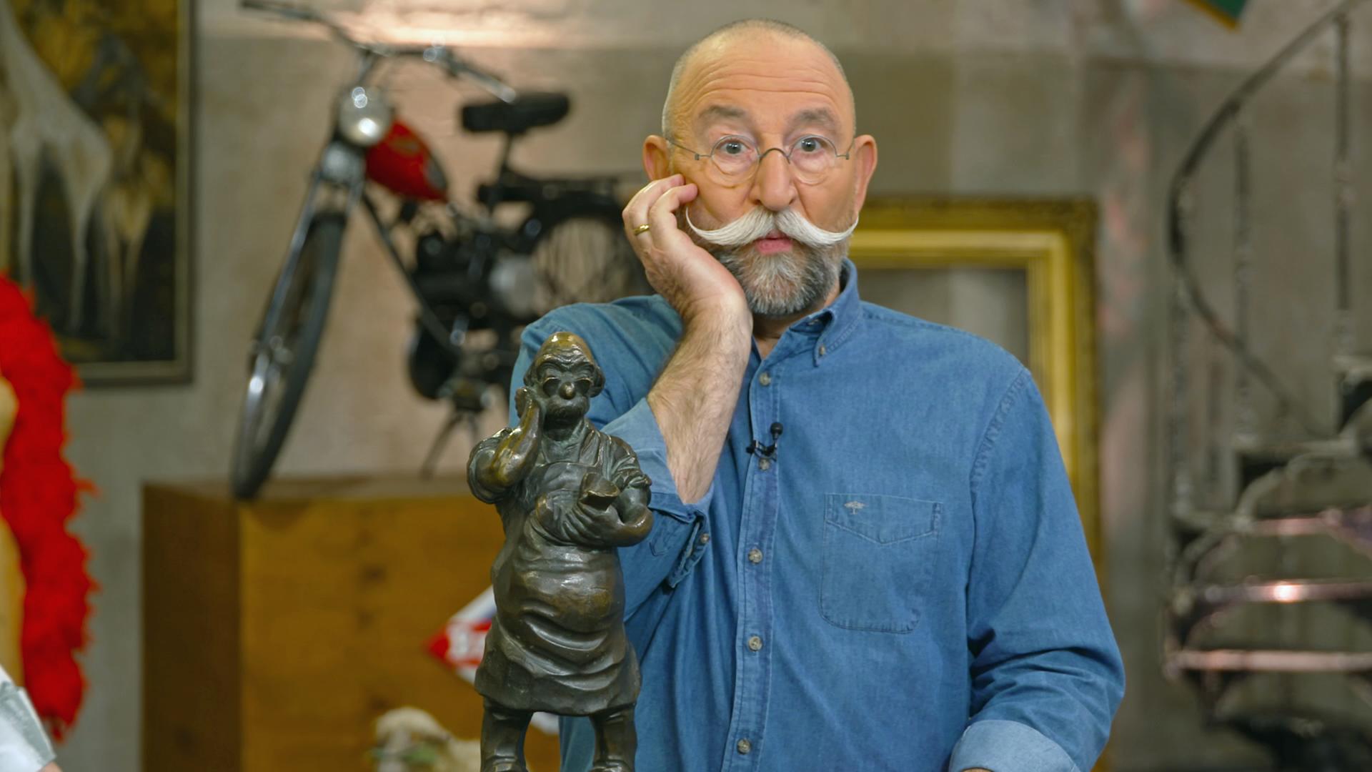 Horst Lichter stellt mit seiner rechten Hand an seiner Wange eine Bronzeskulptur nach, die ihm ähnelt.