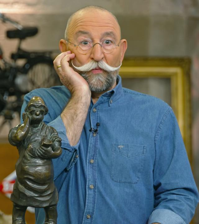 Horst Lichter stellt mit seiner rechten Hand an seiner Wange eine Bronzeskulptur nach, die ihm ähnelt.