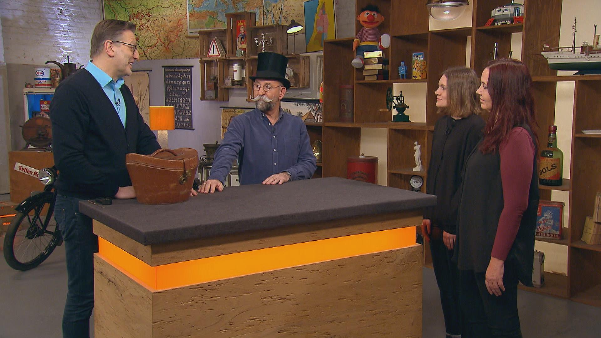 Moderator Horst Lichter steht mit einem Zylinder auf dem Kopf neben dem Experten Detlev Kümmel. Neben dem Expertentisch stehen die Verkäuferinnen.