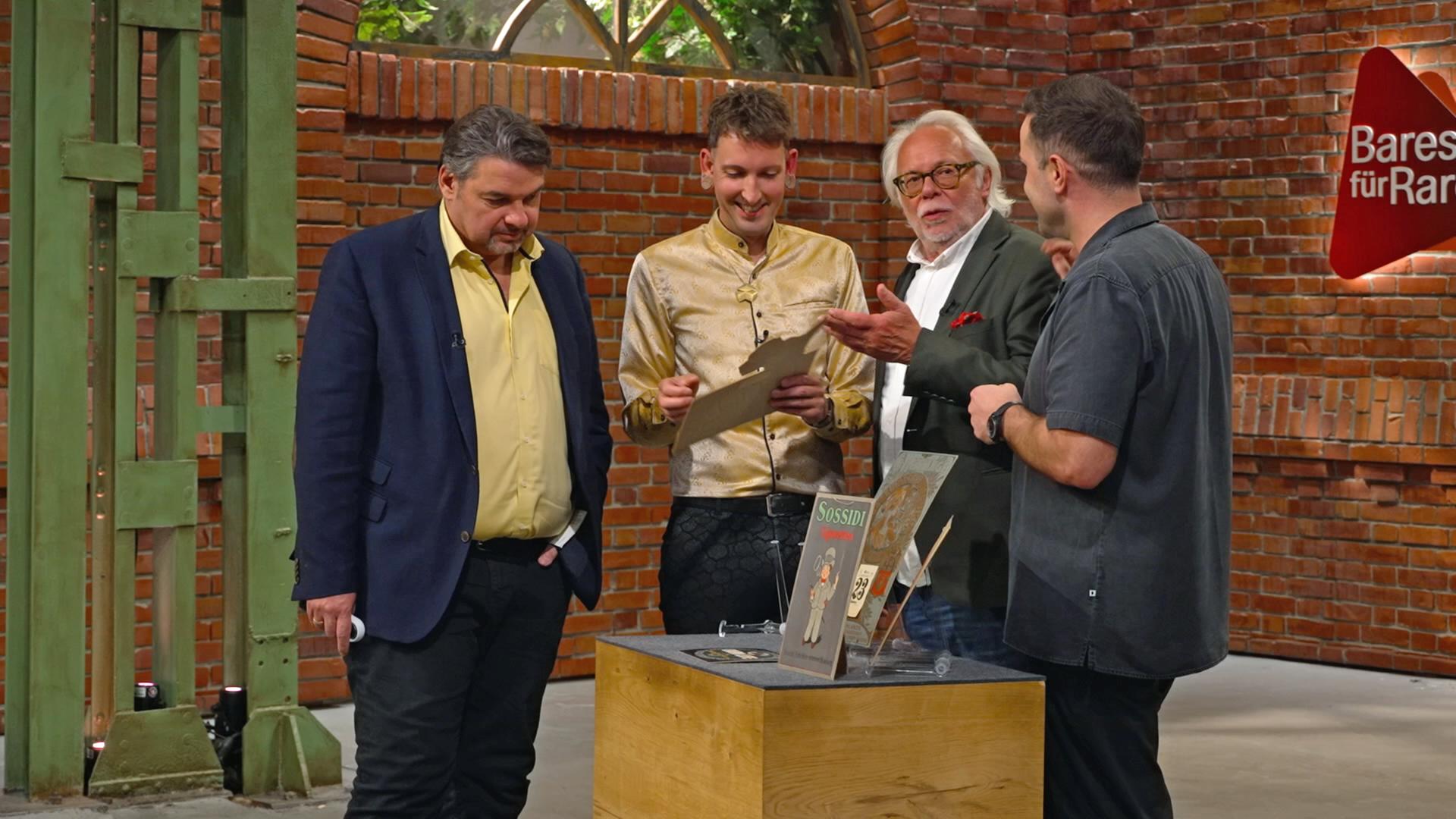 Die Händlerrunde steht um ein Konvolut alter Werbeschilder herum und begutachtet diese genauer.