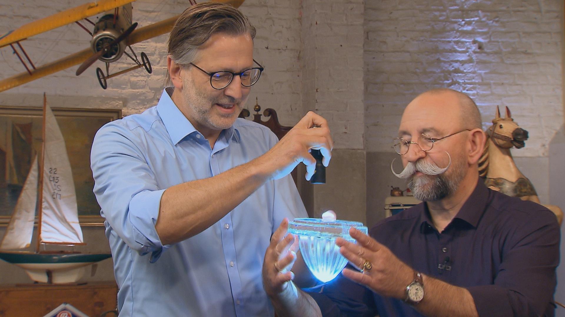 Experte Detlev Kümmel leuchtet mit einer Taschenlampe in ein Glasobjekt, das von Horst Lichter gehalten wird.