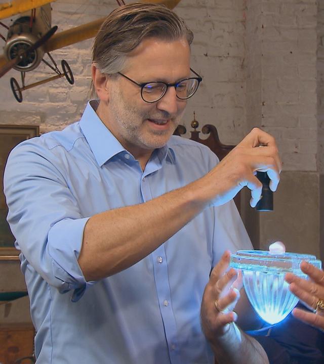 Experte Detlev Kümmel leuchtet mit einer Taschenlampe in ein Glasobjekt, das von Horst Lichter gehalten wird.