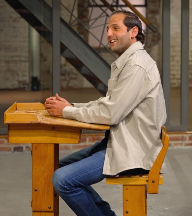 Julian Schmitz-Avila sitzt im Verkaufsraum auf einer alten Schulbank und grinst.