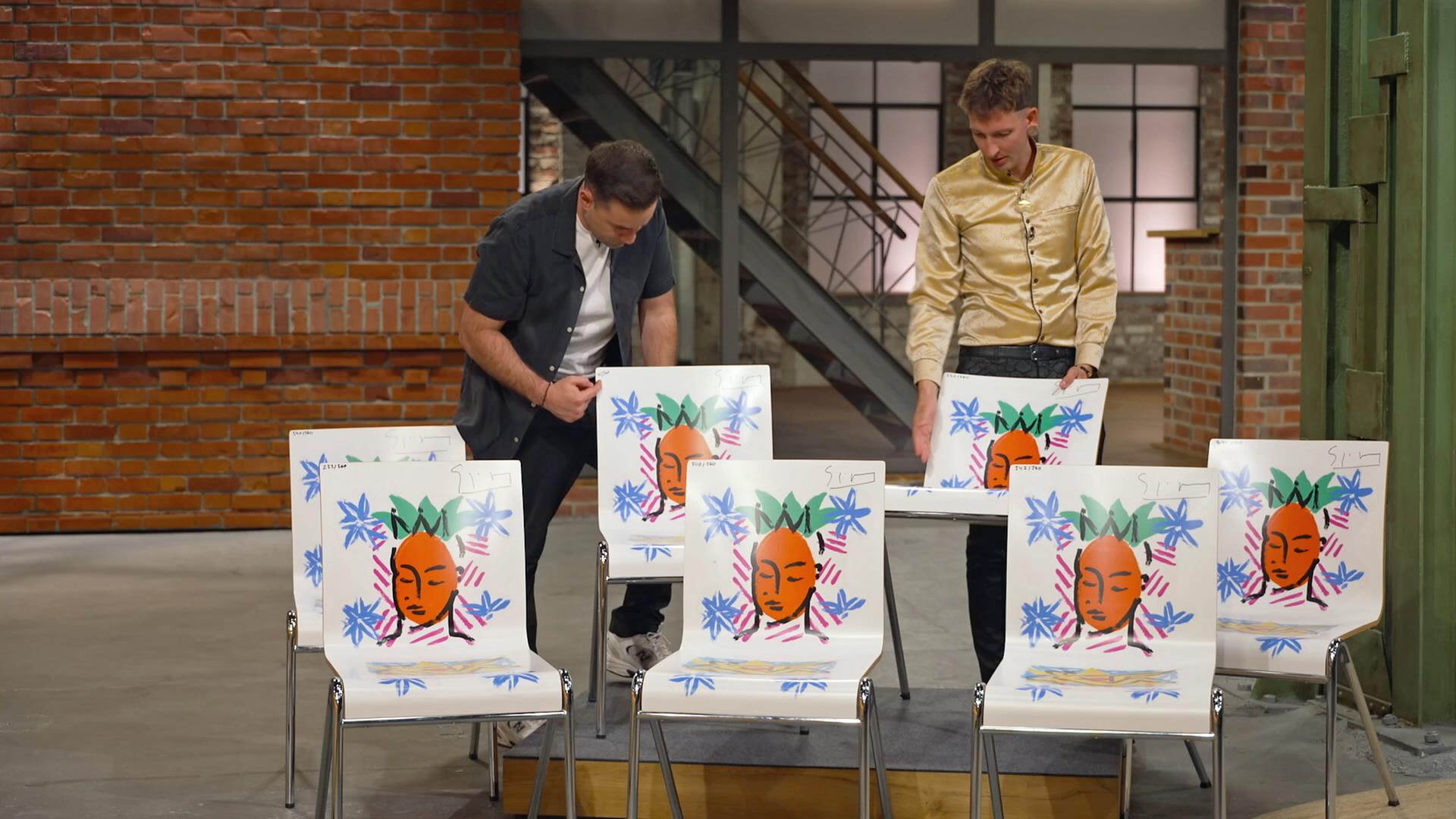 Fabian Kahl und Julian Schmitz-Avila begutachten sieben weiße Stühle mit Motiven genauer. 