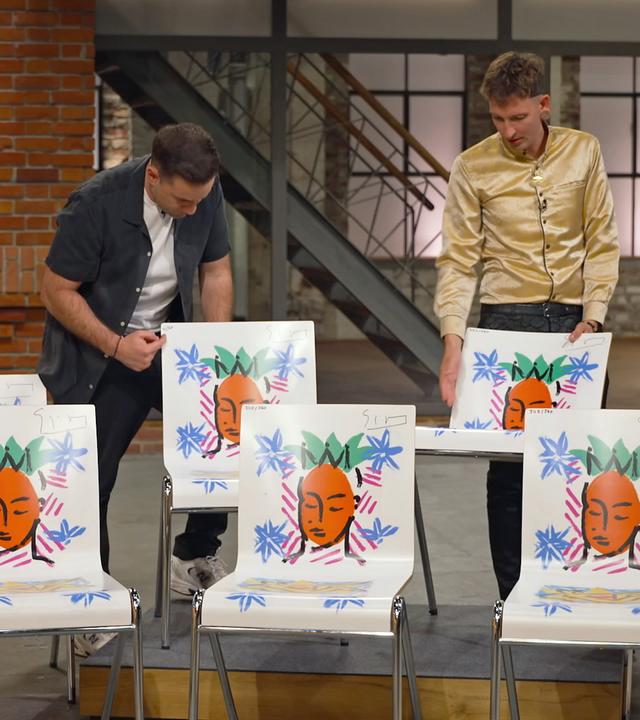 Fabian Kahl und Julian Schmitz-Avila begutachten sieben weiße Stühle mit Motiven genauer. 