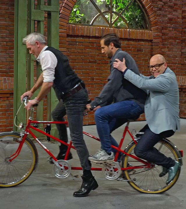 Händler Christian Vechtel sitzt am Steuer eines roten Tandems. David Suppes und Benjamin Leo Leo sitzen hinten mit drauf und sie fahren durch den Händlerraum.