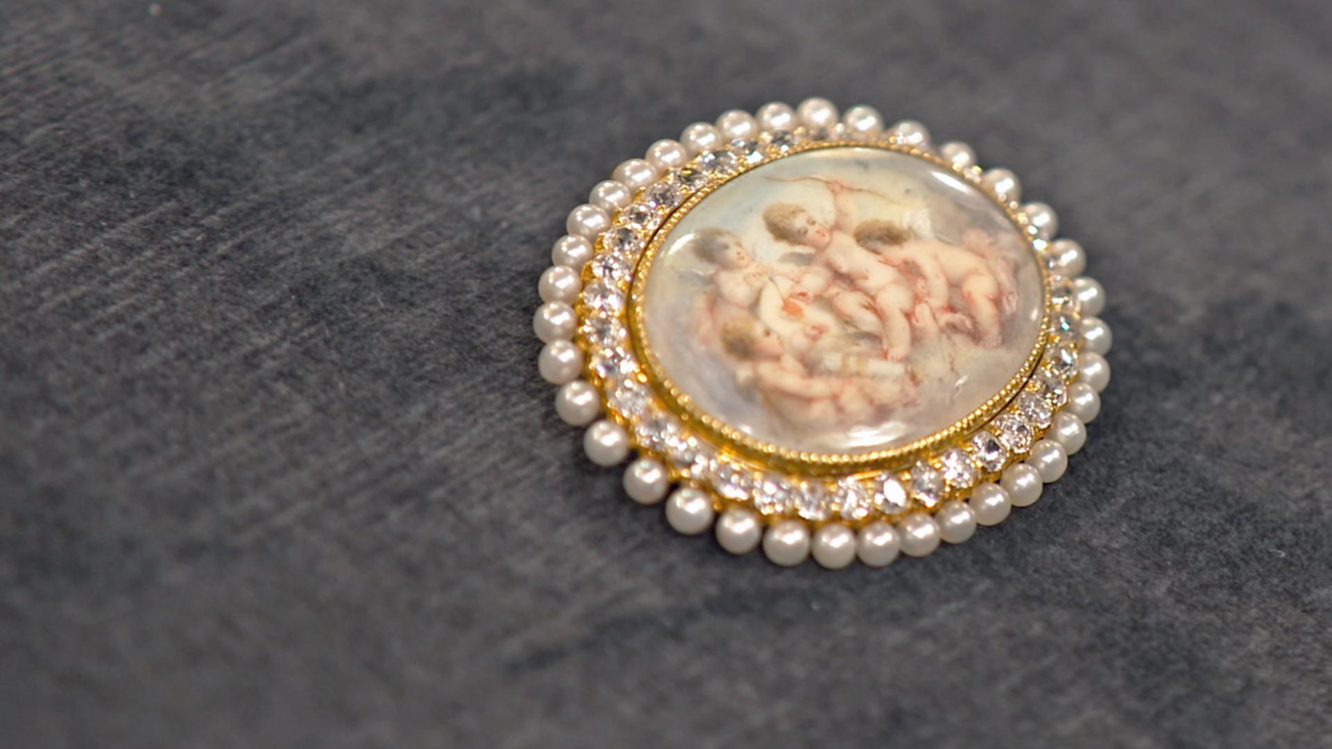 Brosche mit Amor-Motiv umrandet von kleinen Brillanten und Perlen.