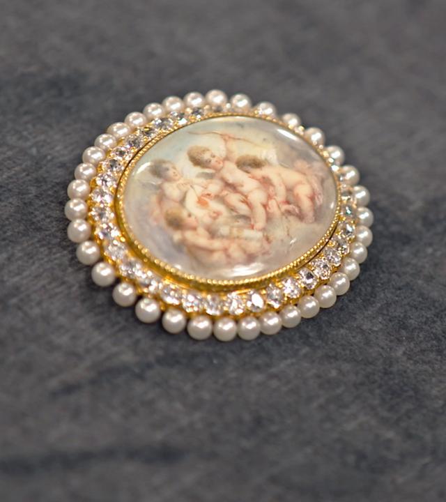 Brosche mit Amor-Motiv umrandet von kleinen Brillanten und Perlen.