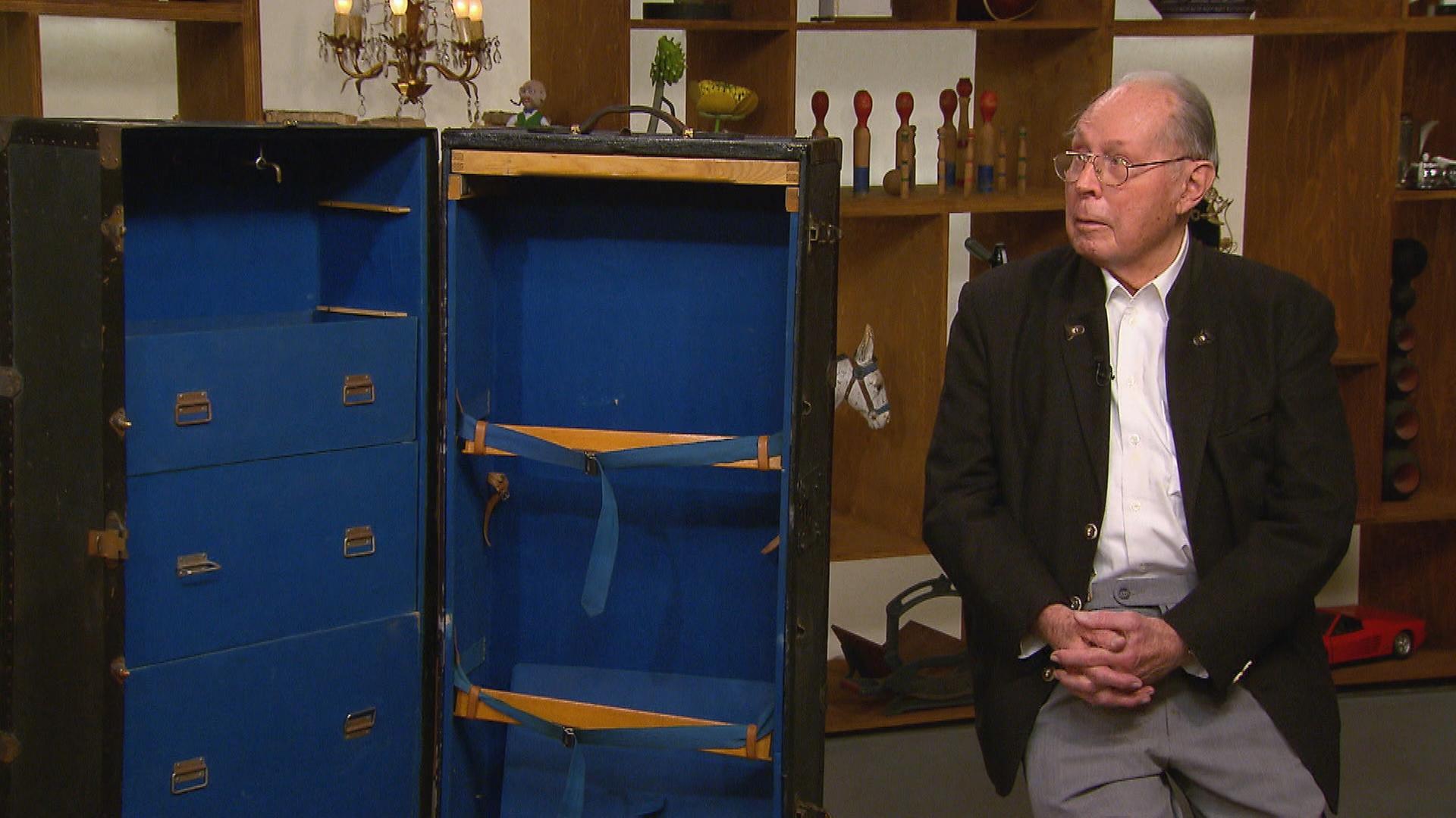 Ein älterer Herr sitzt neben einem großen Schrankkoffer, der innen mit blauem Stoff verkleidet ist.