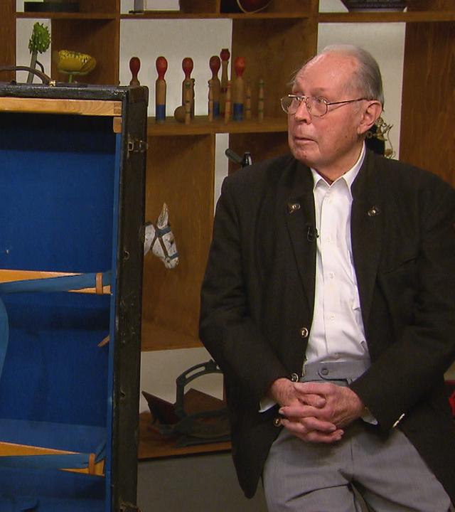 Ein älterer Herr sitzt neben einem großen Schrankkoffer, der innen mit blauem Stoff verkleidet ist.