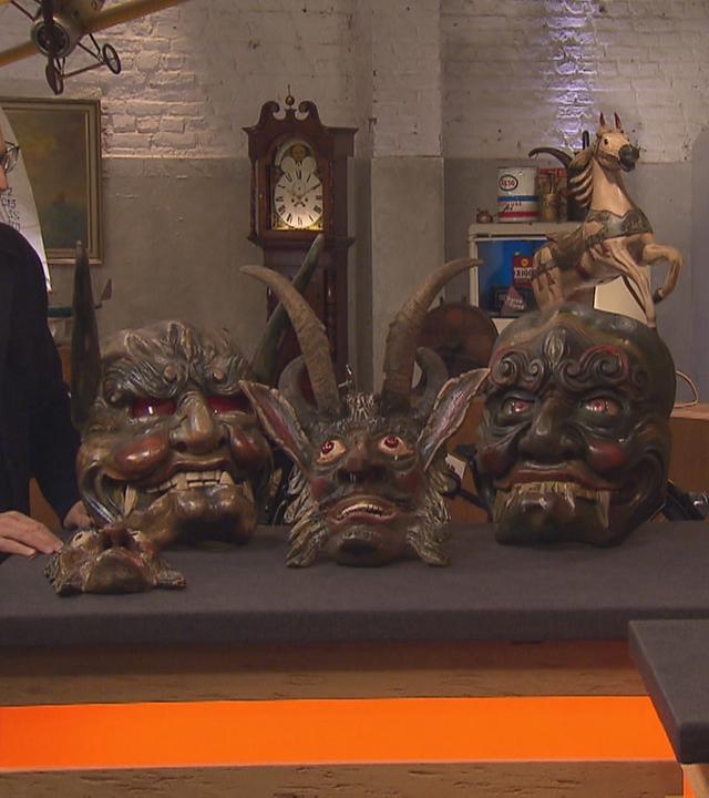 Horst und die Expertise schauen gemeinsam auf die 4 großen Holzmasken zwischen ihnen. Sie haben teilsweise Hörner, grimmige Augen und zeigen ihre Zähne.