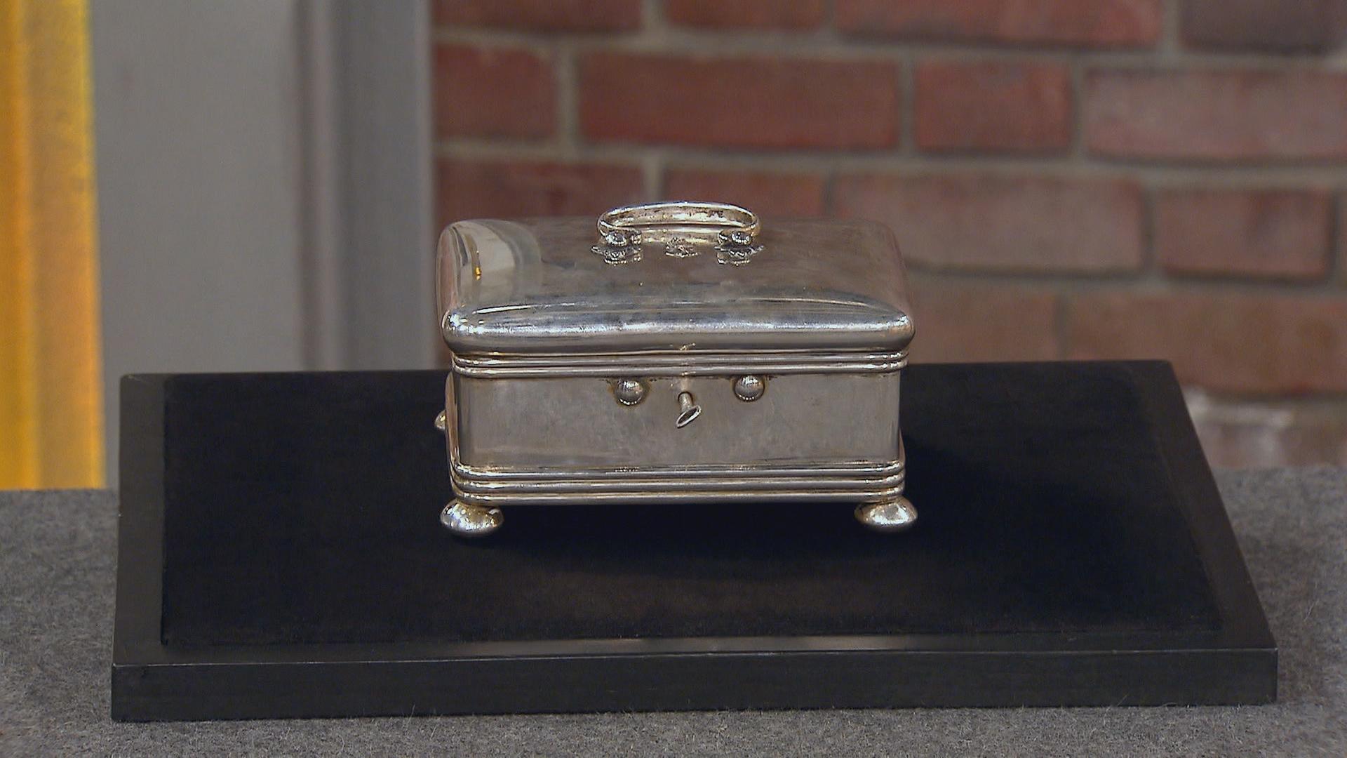 Eine silberne Schatulle steht auf schwarzem Grund. Sie schmückt ein verzierter Griff und kleine Füßchen.