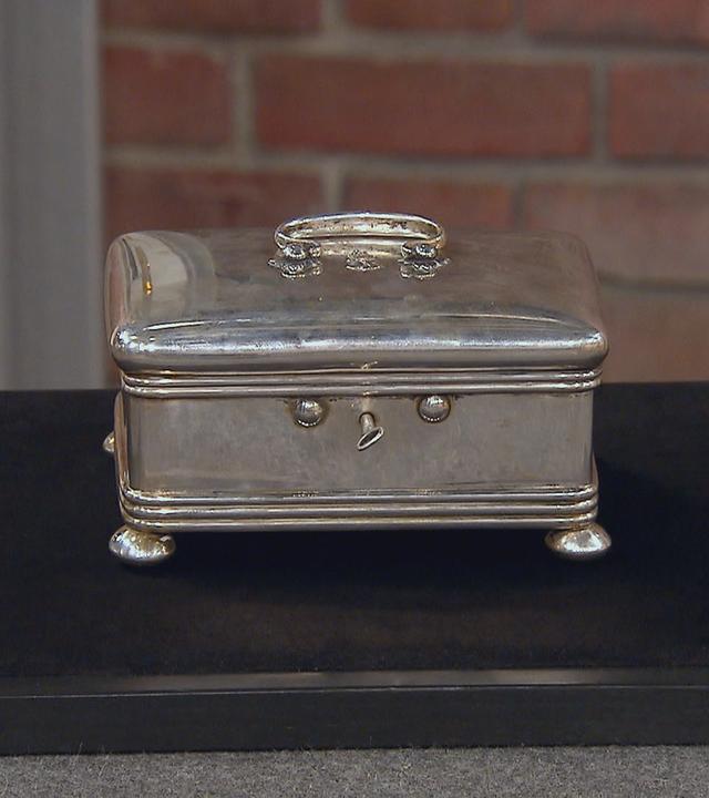 Eine silberne Schatulle steht auf schwarzem Grund. Sie schmückt ein verzierter Griff und kleine Füßchen.