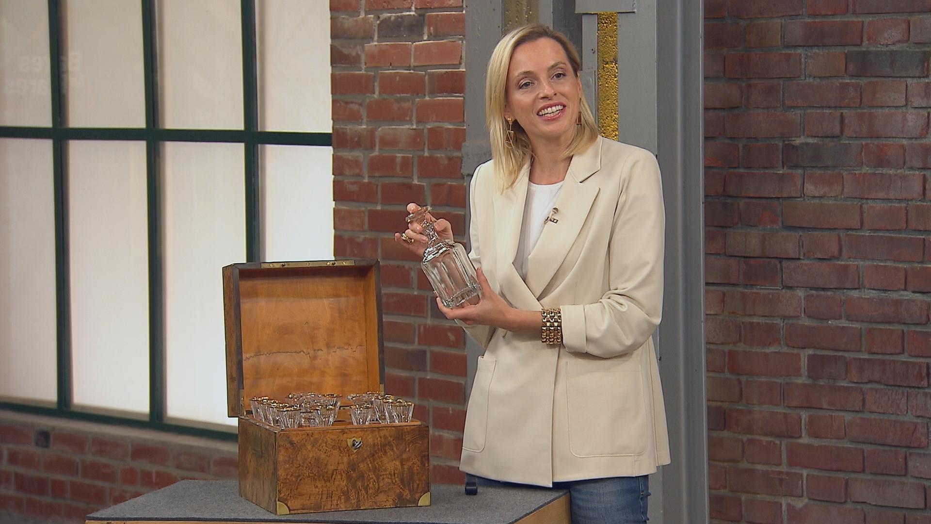 Eine Händlerin hält eine Glasflasche hoch, die sie aus einer Holzkiste mit weiteren Flaschen genommen hat. 