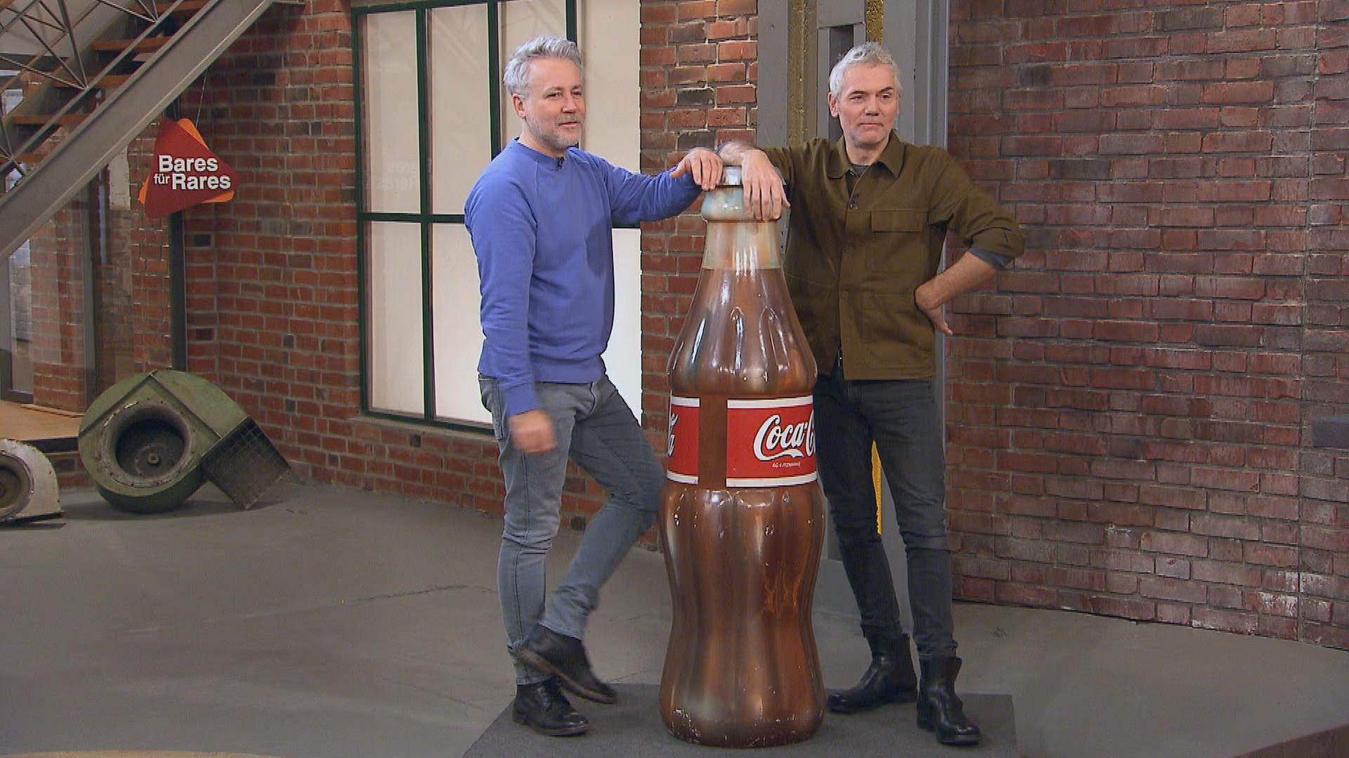 Die Händler Jan Cizek und Christian Vechtel stehen neben einer riesigen Coca-Cola-Flasche