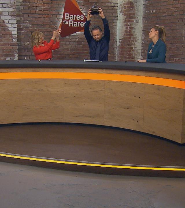 Walter „Waldi“ Lehnertz, Elke Velten, Wolfgang Pauritsch, Sarah Schreiber und Jos van Katwijk sitzen am Händlerpult. Wolfgang Pauritsch hält eine Kupferdose in die Luft.