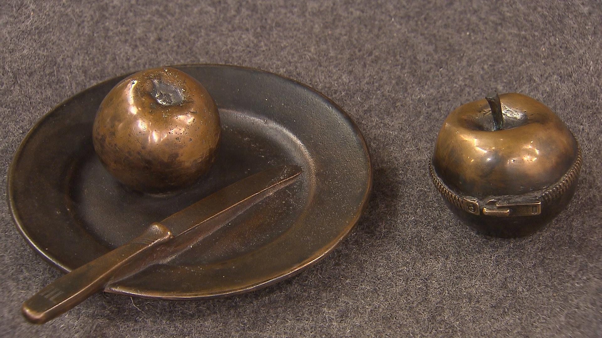 Zwei kleine Bronzeskulpturen. Links ein Apfel auf einem Teller mit Messer, rechts ein Apfel mit Reißverschluss.