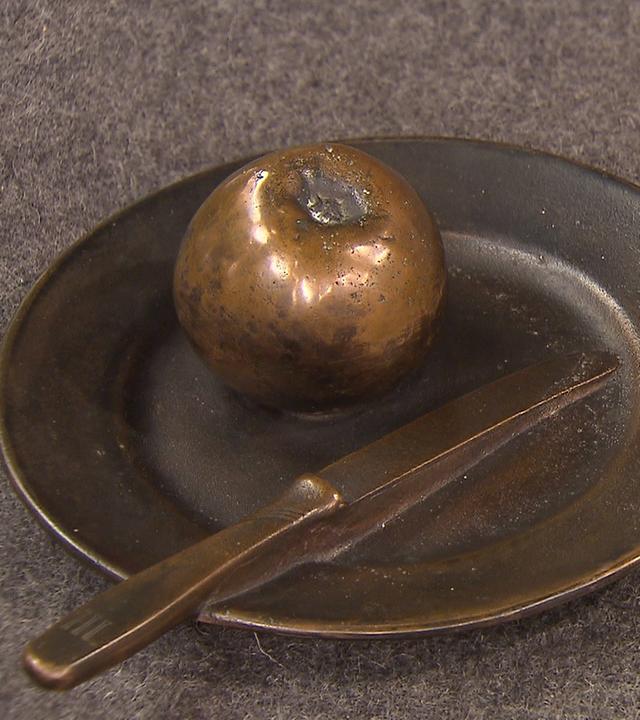 Zwei kleine Bronzeskulpturen. Links ein Apfel auf einem Teller mit Messer, rechts ein Apfel mit Reißverschluss.