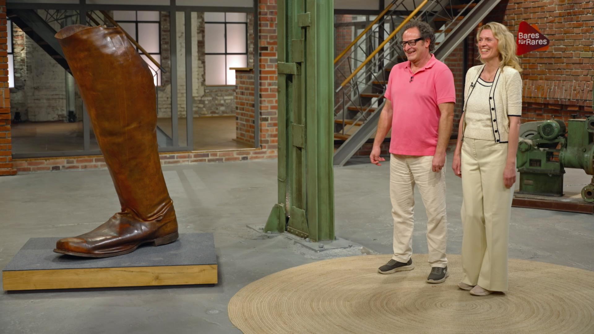 Im Händlerraum steht rechts ein Verkäuferpaar und strahlt. Links steht das Objekt: Ein überdimensionaler, brauner Lederstiefel.
