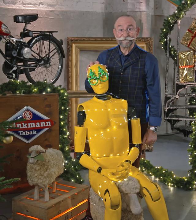 Horst Lichter steht hinter einem sitzenden, gelben Roboter. Der Roboter trägt eine grüne, glitzernde Brille mit Tannenmotiv.
