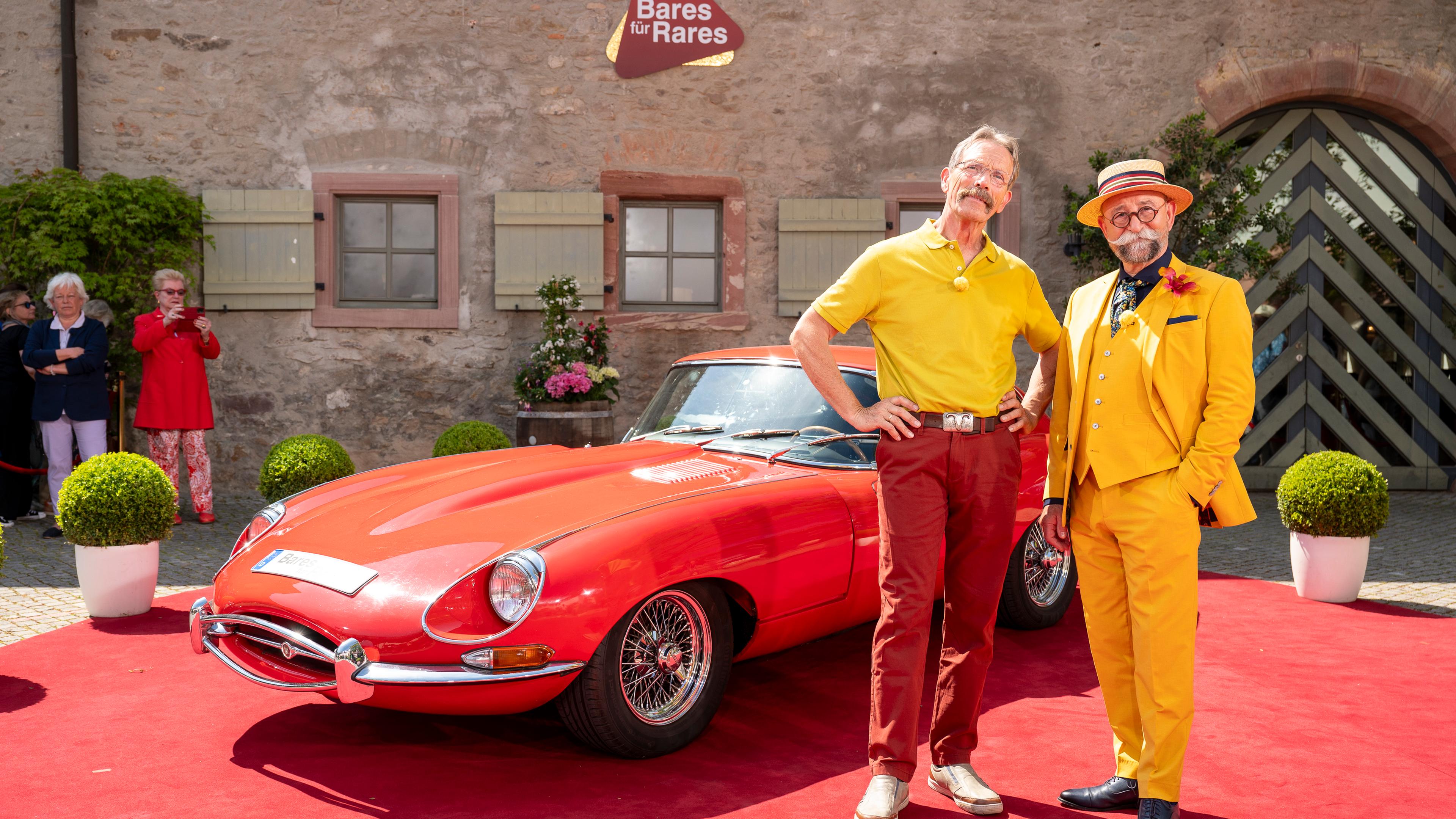 Klaus Thiedig und Horst Lichter stehen auf einem roten Teppich und lächeln in die Kamera. Hinter ihnen ist ein roter Jaguar E-Type Coupé, Baujahr 1968 zu sehen.