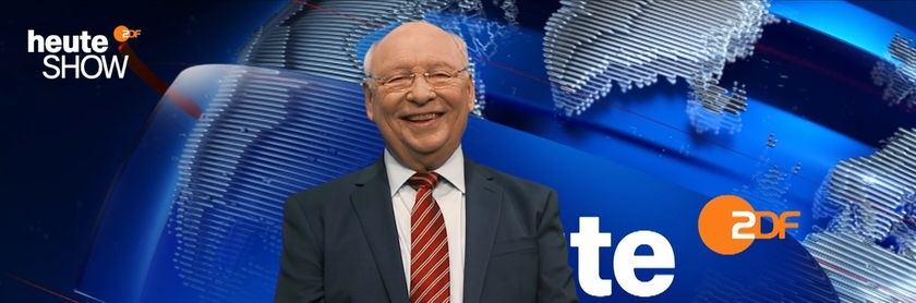 Vor dem blau leuchtenden Hintergrund der „heute-show“ lächelt Gernot Hassknecht in die Kamera. Der glatzköpfige Mann mit Brille trägt einen dunklen Anzug und eine rote Streifenkrawatte. Oben links sind der Schriftzug der Sendung und rechts das ZDF-Logo zu sehen.