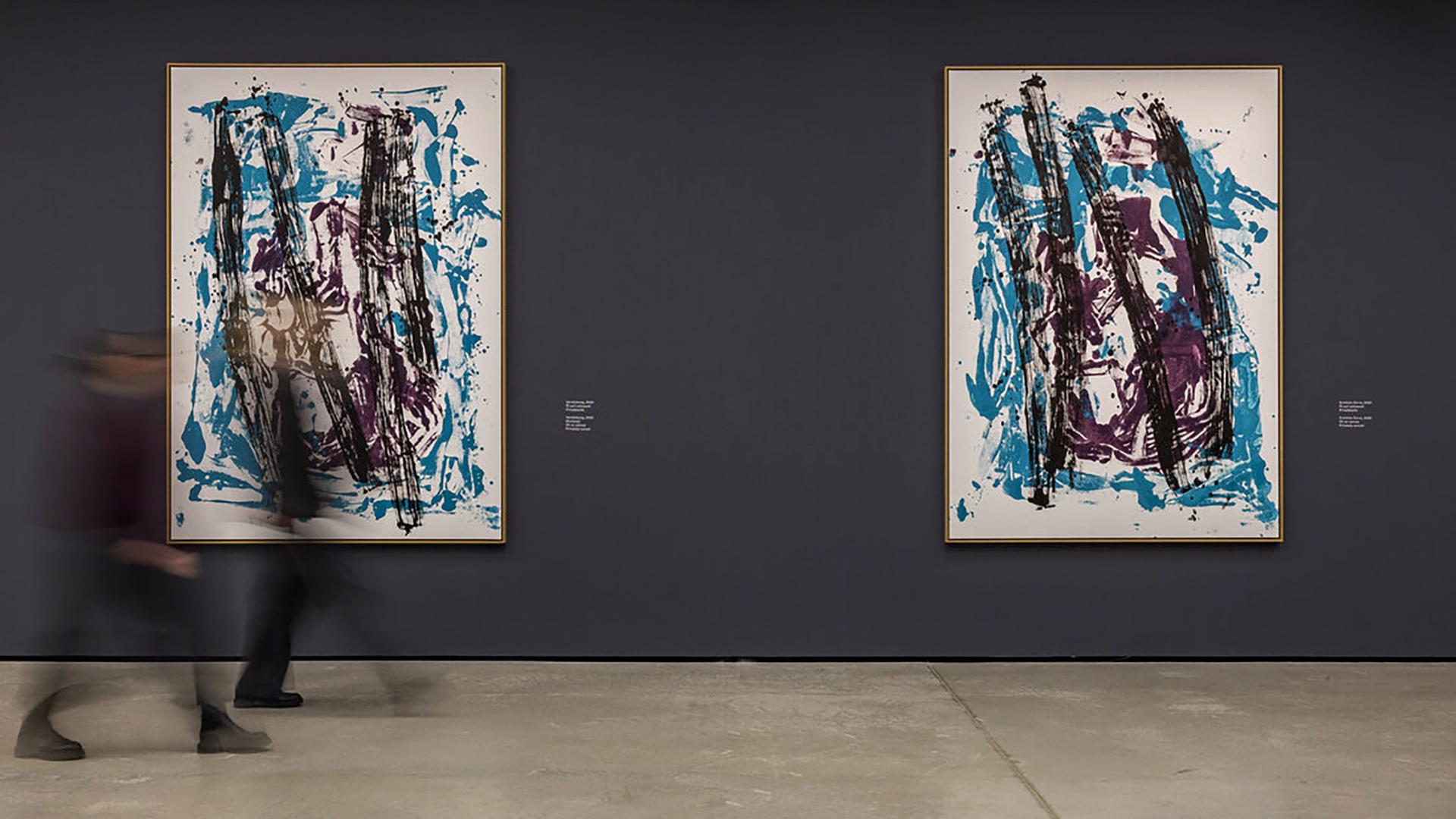 Foto von zwei großen abstrakten Drucken, die an einer grauen Wand hängen. Davor sind zwei in der Bewegung verwischte Menschen zu sehen.