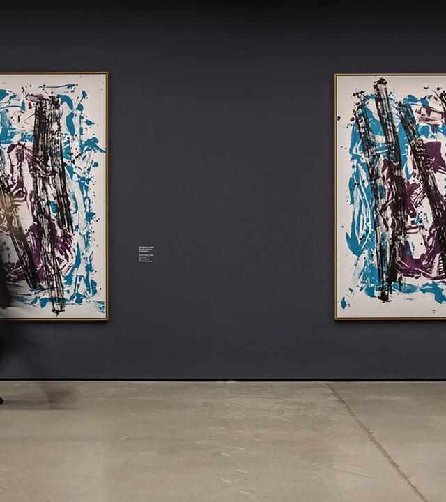 Foto von zwei großen abstrakten Drucken, die an einer grauen Wand hängen. Davor sind zwei in der Bewegung verwischte Menschen zu sehen.