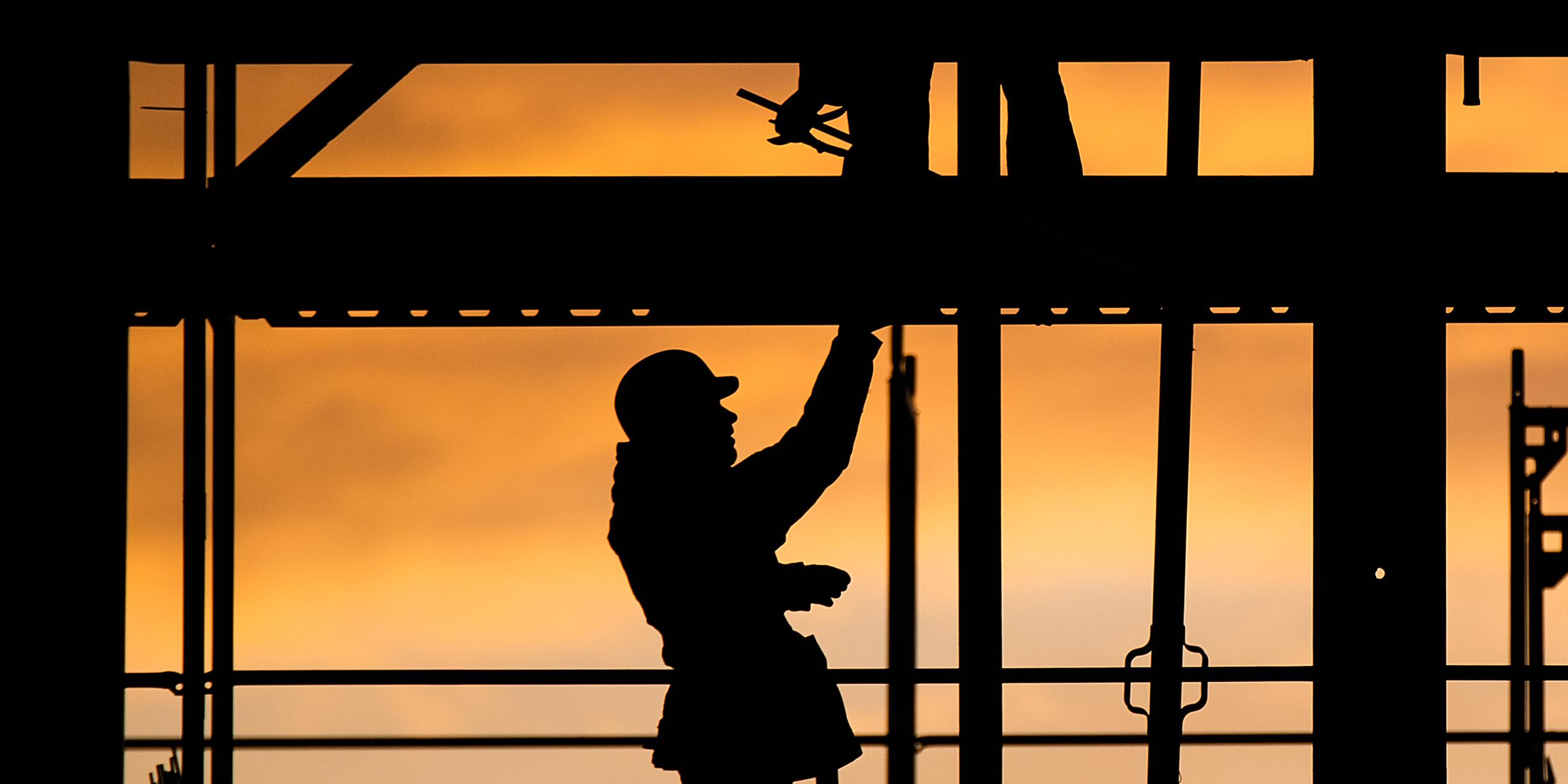 Die Silhouette eines Bauarbeiters zeichnet sich auf einer Baustelle vor dem verfärbten Morgenhimmel ab. 
