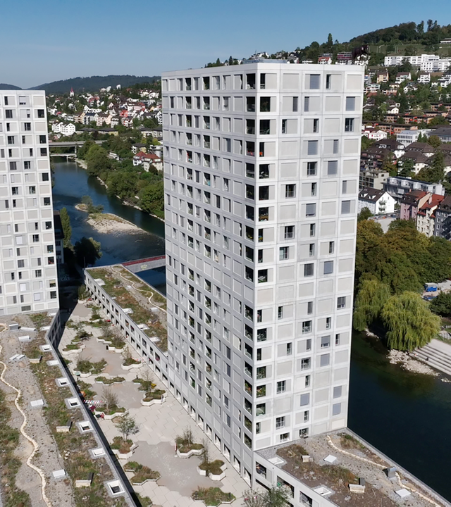 Das Bild zeigt zwei hohe, moderne Wohngebäude mit einer strukturierten Fassade. Im Vordergrund verläuft eine Straße, während im Hintergrund Bäume und weitere Gebäude zu sehen sind. Ein Fluss fließt neben den Gebäuden.