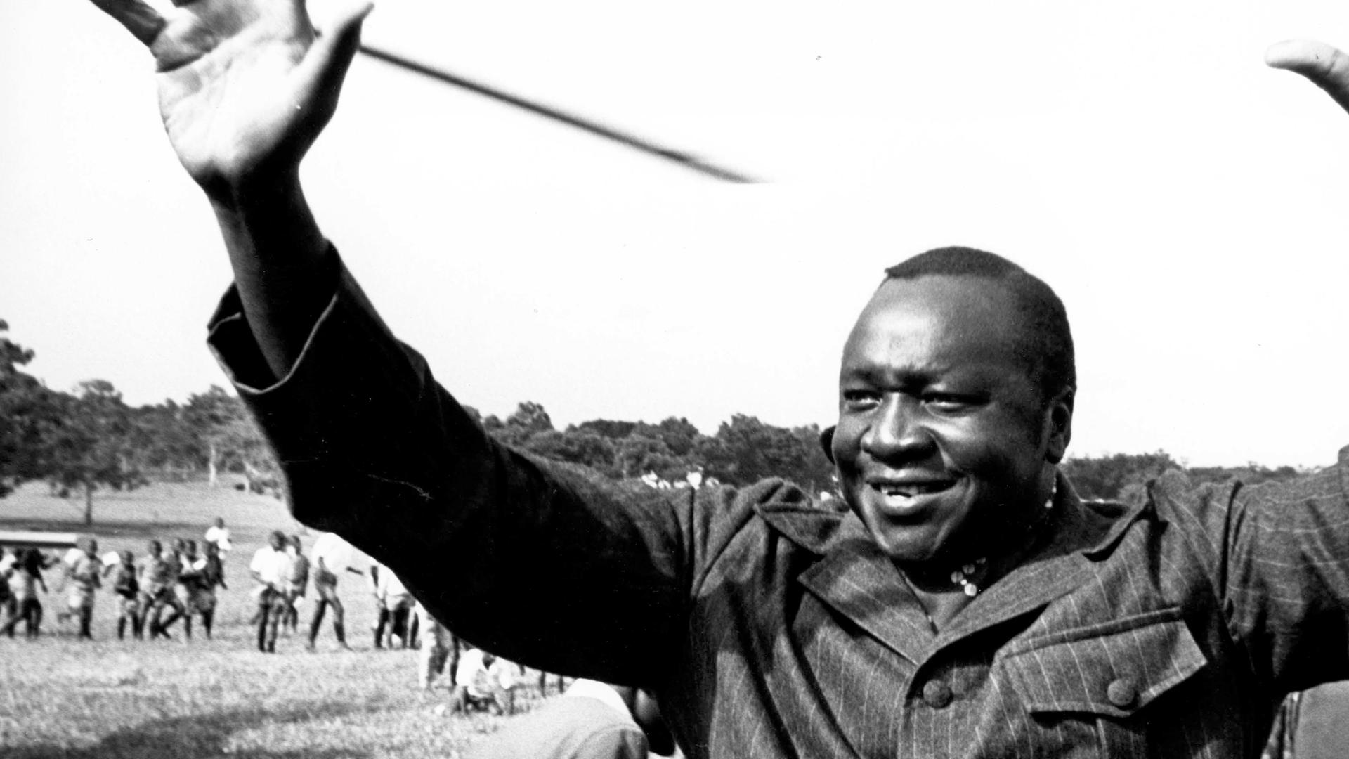 "Bauplan des Bösen: Idi Amin": Schwarz-Weiß-Aufnahme: Ein Mann hat beide Arme erhoben, sodass die Handflächen frontal zu sehen sind. Er lächelt. Im Hintergrund viele versammelte Menschen.