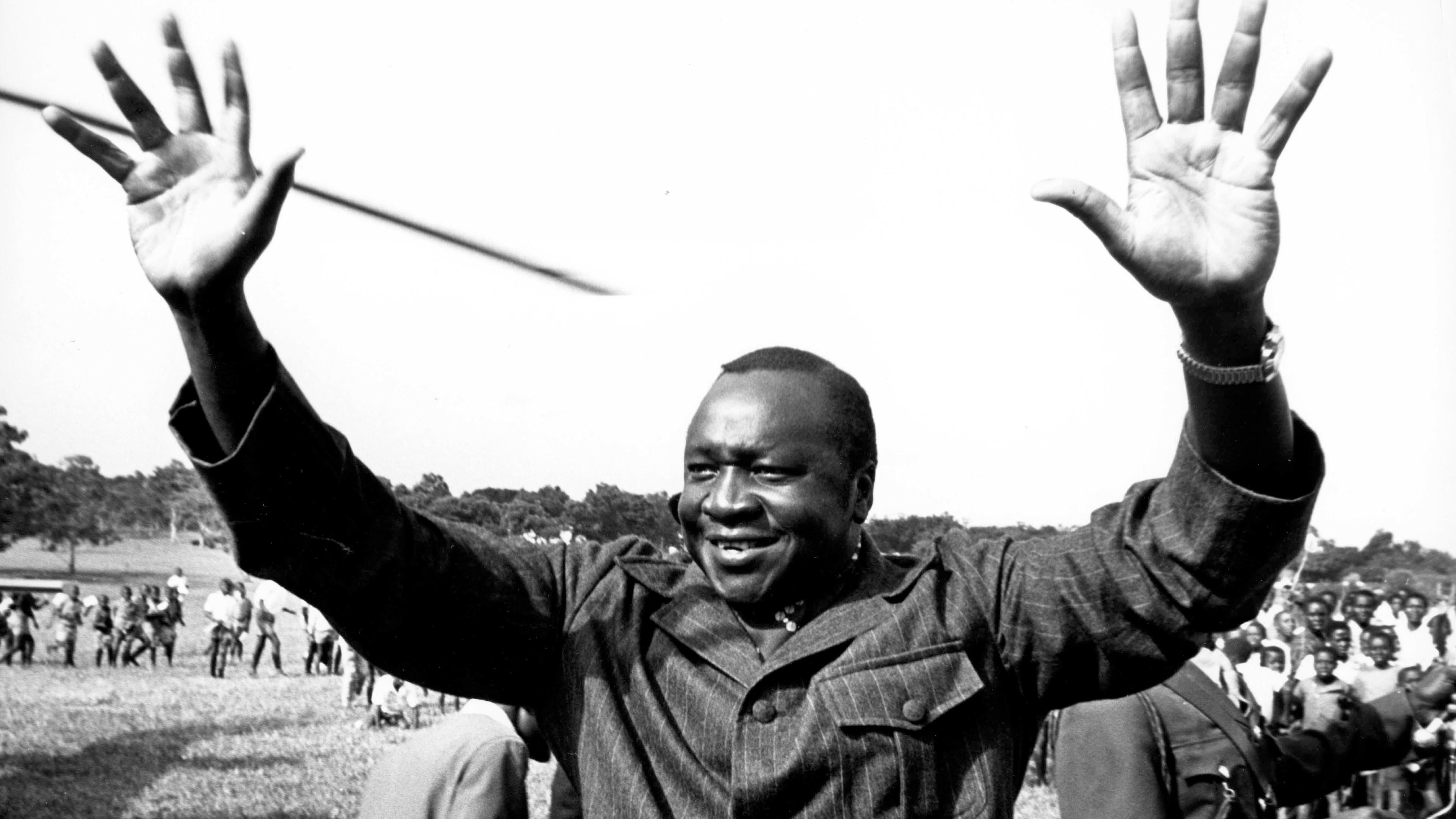 "Bauplan des Bösen: Idi Amin": Schwarz-Weiß-Aufnahme: Ein Mann hat beide Arme erhoben, sodass die Handflächen frontal zu sehen sind. Er lächelt. Im Hintergrund viele versammelt Menschen.