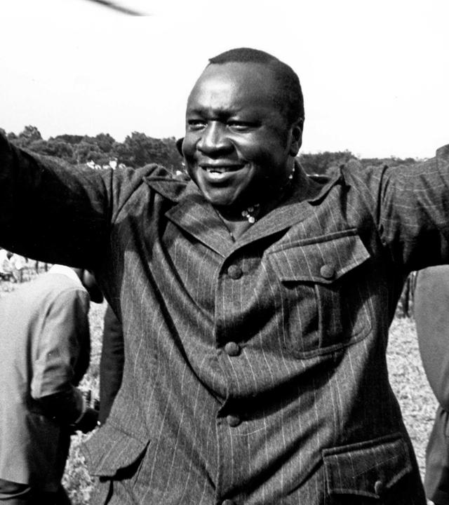 "Bauplan des Bösen: Idi Amin": Schwarz-Weiß-Aufnahme: Ein Mann hat beide Arme erhoben, sodass die Handflächen frontal zu sehen sind. Er lächelt. Im Hintergrund viele versammelte Menschen.
