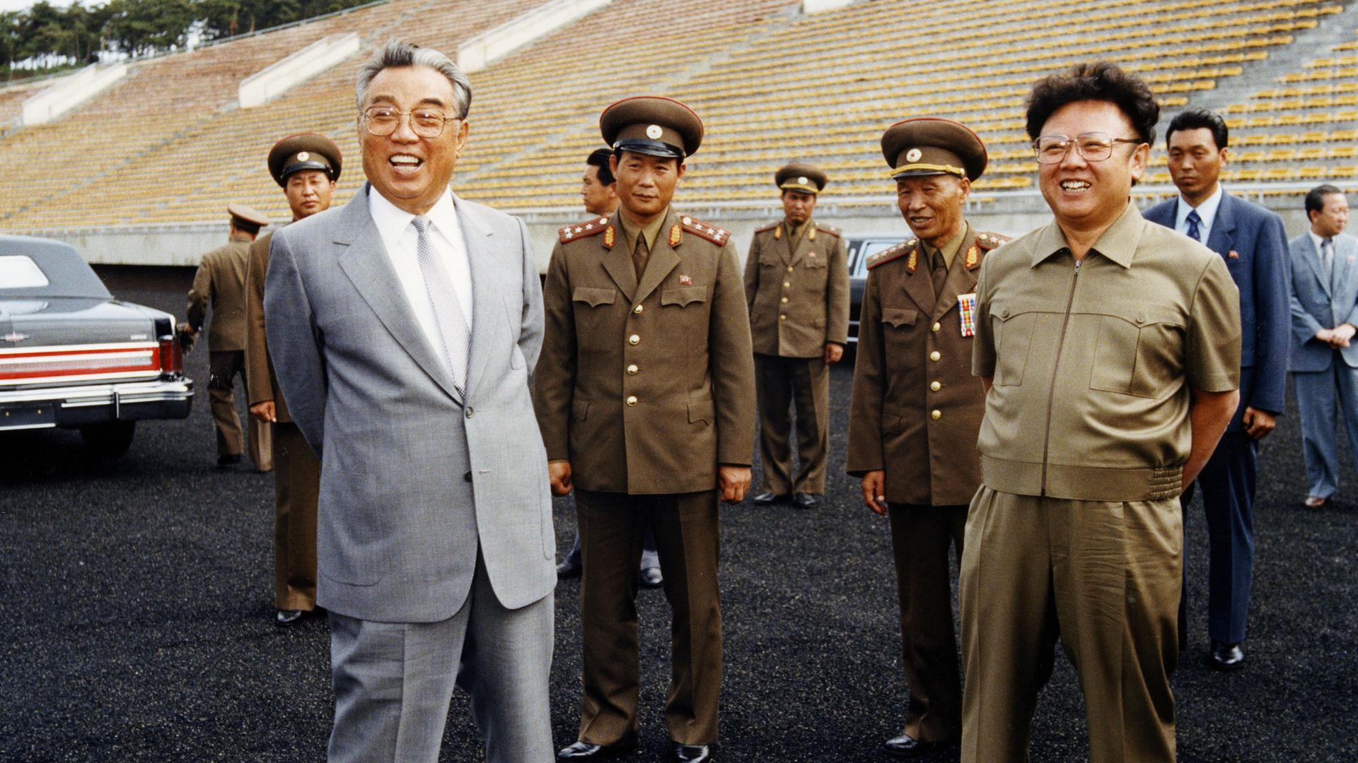 "Bauplan des Bösen: Kim Il-Sung": Ein älterer Mann steht in einem Stadion, hinter ihm leere Ränge. Er trägt eine Brille, einen grauen Anzug mit hellgrauer Krawatte und lacht. Hinter ihm stehen uniformierte Männer.