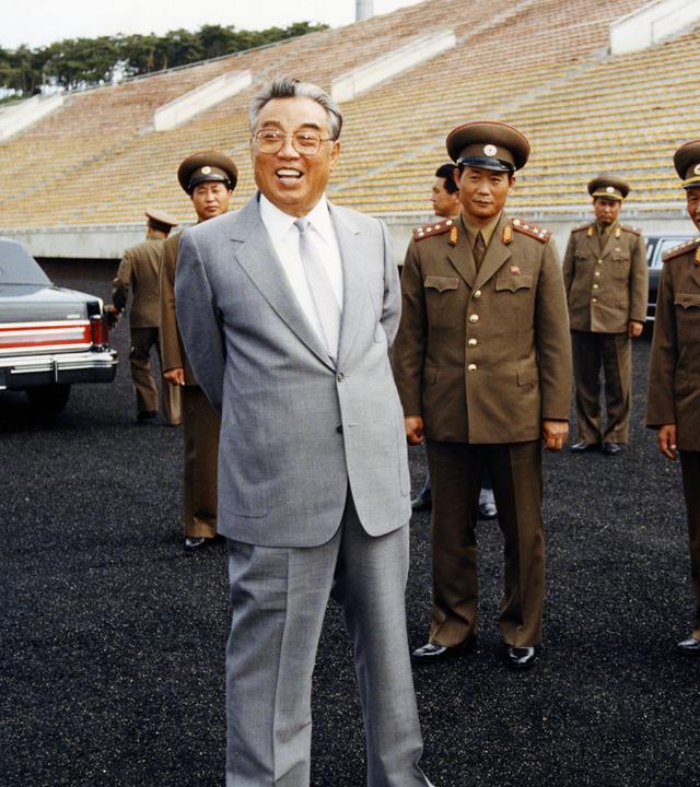 "Bauplan des Bösen: Kim Il-Sung": Ein älterer Mann steht in einem Stadion, hinter ihm leere Ränge. Er trägt eine Brille, einen grauen Anzug mit hellgrauer Krawatte und lacht. Hinter ihm stehen uniformierte Männer.
