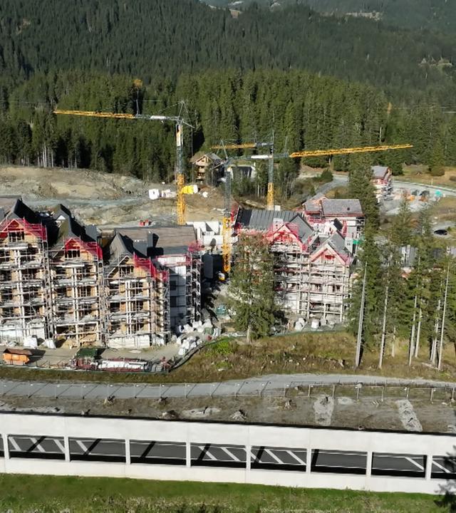 Das Bild zeigt eine Baustelle in den Alpen, konkret am Pass Thurn. Im Vordergrund sind unvollendete Gebäude eines Luxusresorts zu sehen, die von Gerüsten umgeben sind. Einige Teile der Fassaden sind bereits sichtbar, während andere noch Rohbauzustand haben. In der Umgebung sind Kräne aufgestellt, die für den Bau verwendet werden.   Der Untergrund ist teilweise unebenen und es gibt Stellen mit Schotter. Die Landschaft im Hintergrund ist von dichten Wäldern geprägt, die sich bis zu den Berggipfeln erstrecken. Auf der linken Seite des Bildes ist eine Straße sichtbar, die entlang der Baustelle verläuft. Diese Umgebung vermittelt den Eindruck eines stillgelegten, unfertigen Projekts, das sich gegen die natürliche Kulisse der Alpen abhebt.