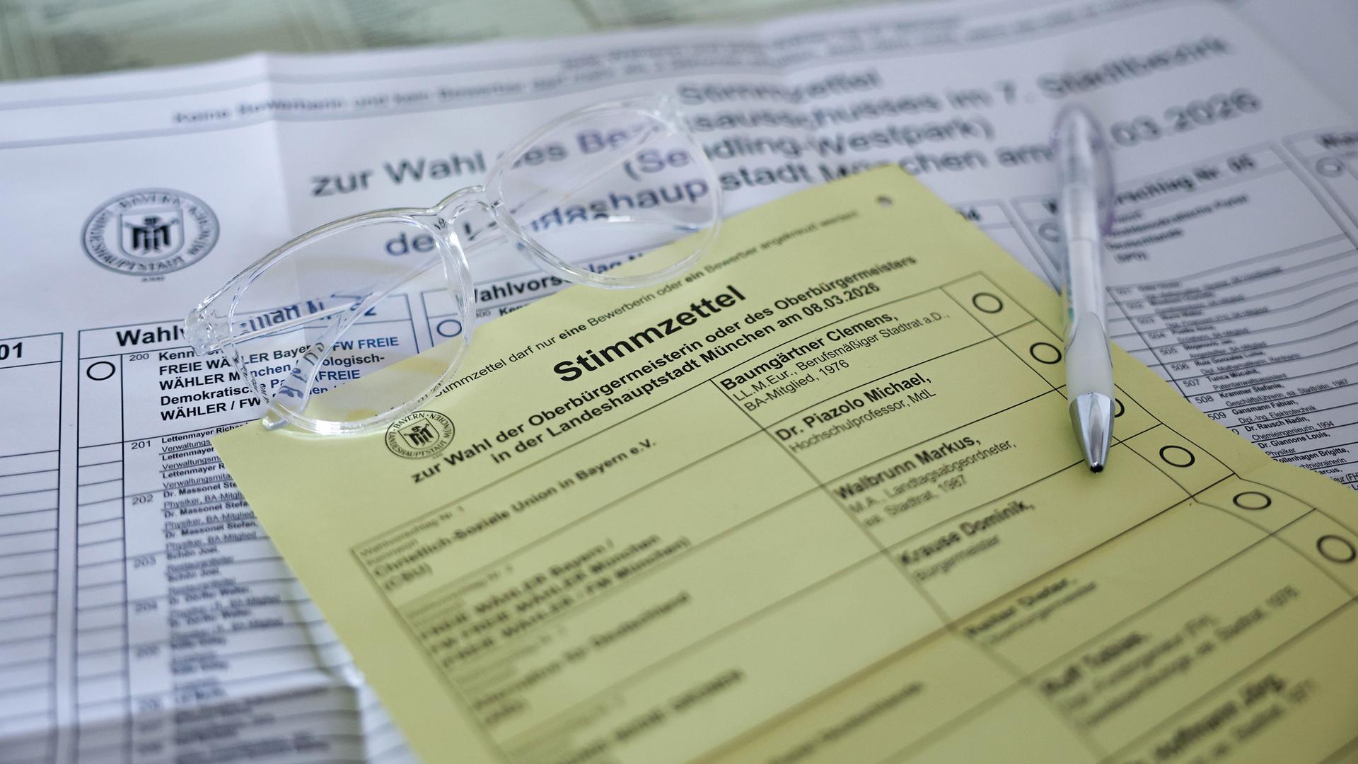 Briefwahlunterlagen für die Kommunalwahl in Bayern 2026 liegen auf dem Tisch. Darauf liegt eine Brille mit durchsichtigem Rahmen.