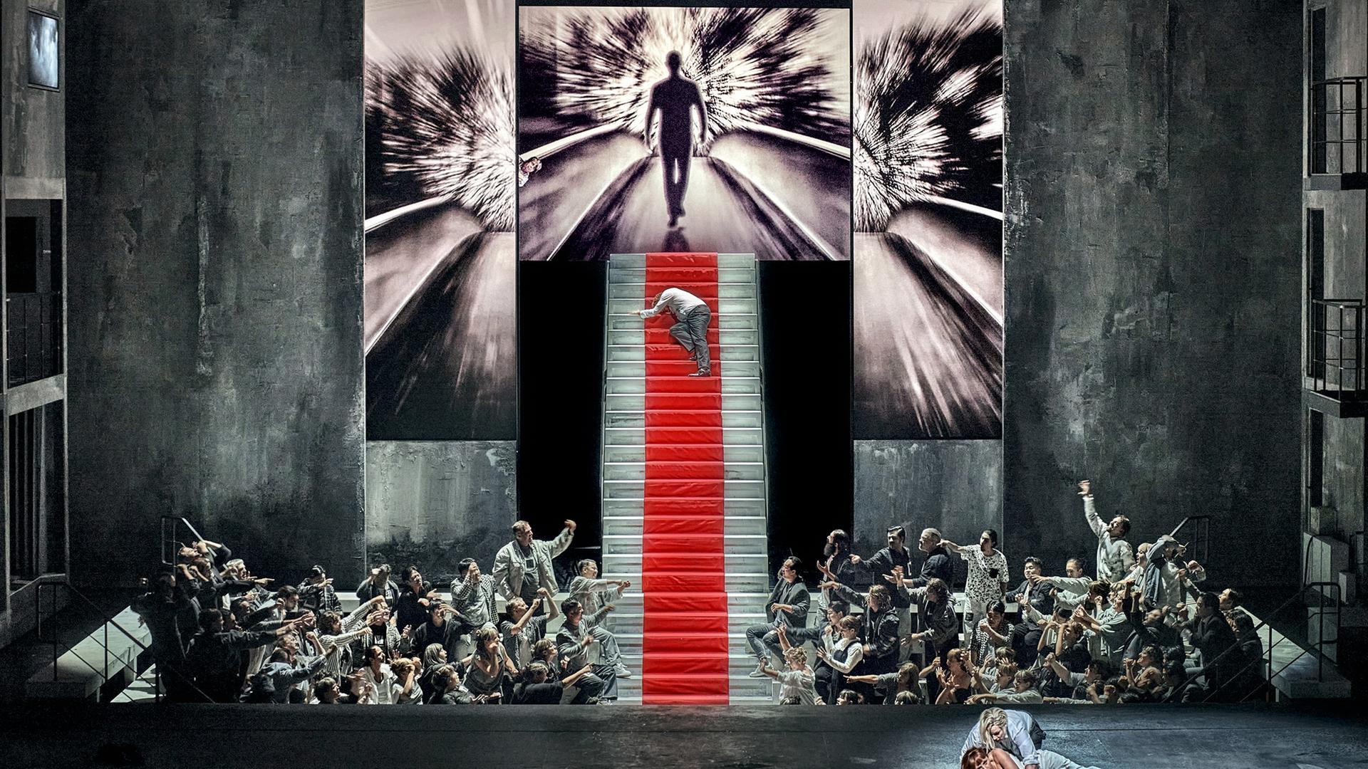 Bühne mit steiler Treppe und zahlreichen Akteuren, die zur Treppe hinweisen, auf der ein Mensch hochkrabbelt
