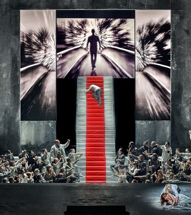 Bühne mit steiler Treppe und zahlreichen Akteuren, die zur Treppe hinweisen, auf der ein Mensch hochkrabbelt