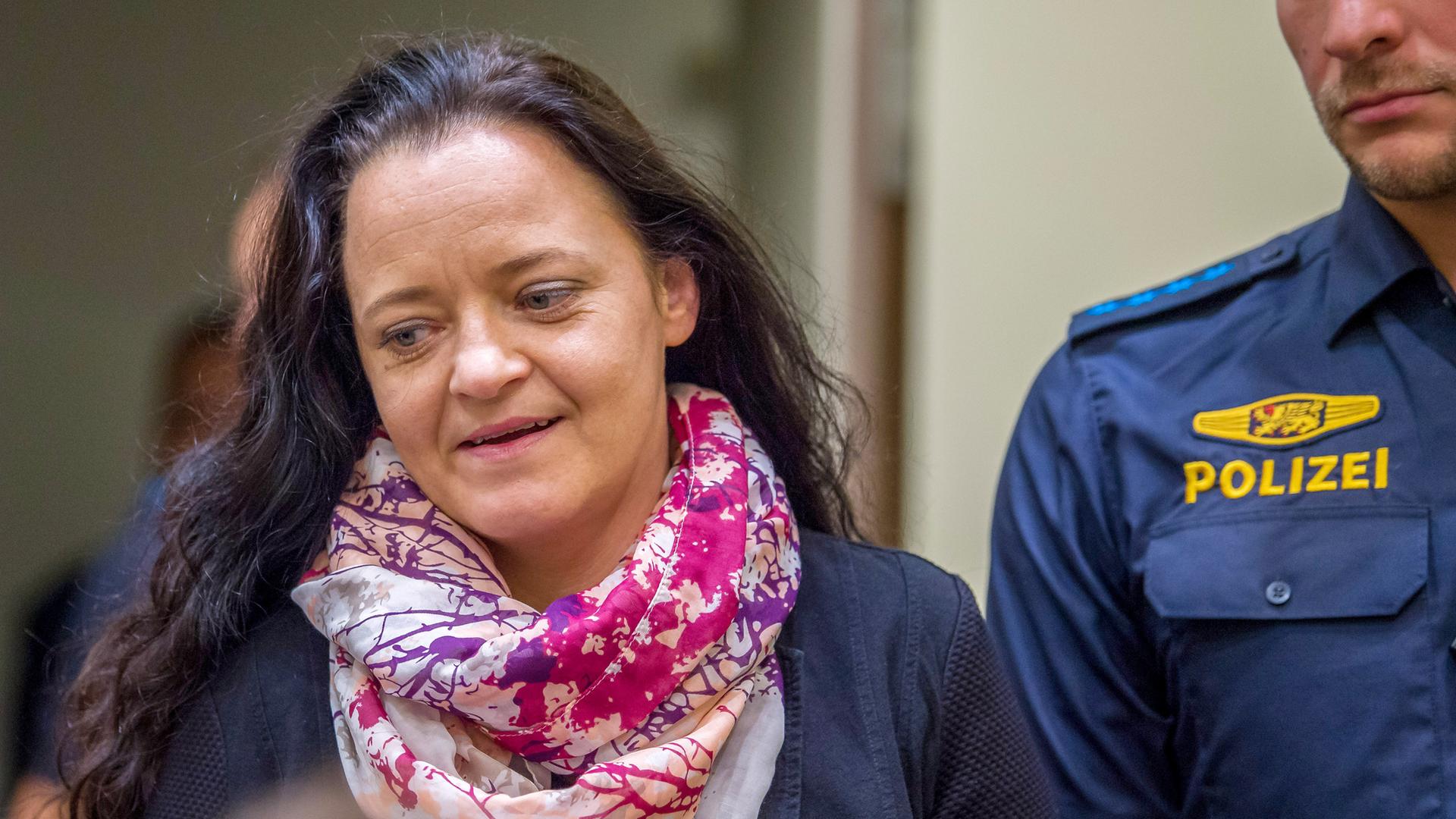 Eine Frau mittleren Alters mit langen Haaren und buntem Schal wird von einem Polizisten begleitet.