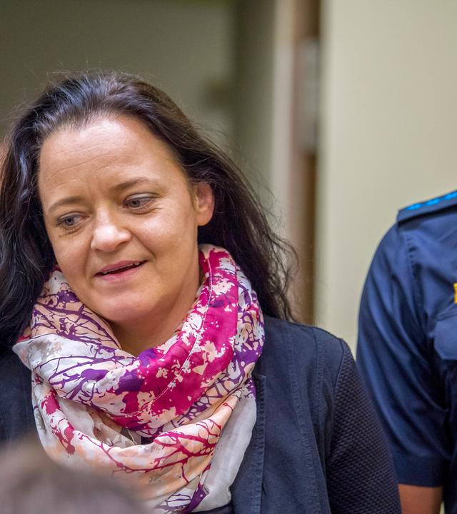 Eine Frau mittleren Alters mit langen Haaren und buntem Schal wird von einem Polizisten begleitet.