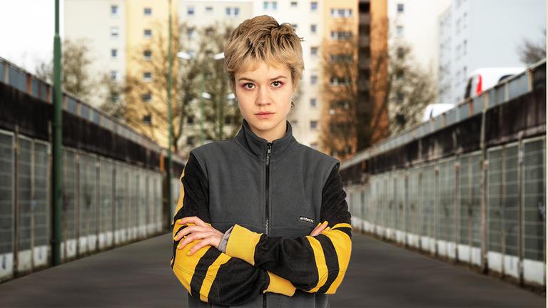 "Becoming Charlie": Charlie (Lea Drinda) steht mit verschränkten Armen zwischen zwei Parkgaragen und blickt in die Kamera. Im Hintergrund ein Plattenbau-Hochhaus.